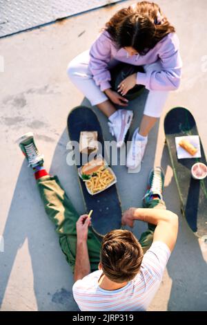 Draufsicht auf Jugendliche, die auf ihren Skateboards Fast Food essen. Selektiver Fokus auf den Kopf eines Teenagers, der mit seiner Freundin sitzt. Stockfoto