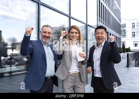 Erfolgreiches, vielfältiges Business-Team, drei liebevolle Männer und Frauen, die die Kamera betrachten und glücklich den Sieg feiern, Team träumen vor dem Bürogebäude, Business Group in Business-Anzügen Stockfoto