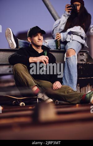 Ein junger Skater-Junge und seine Freundin sitzen auf dem Dach des Gebäudes, trinken Bier und haben Spaß am Abend. Stockfoto