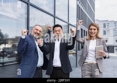 Erfolgreiches, vielfältiges Business-Team, drei liebevolle Männer und Frauen, die die Kamera betrachten und glücklich den Sieg feiern, Team träumen vor dem Bürogebäude, Business Group in Business-Anzügen Stockfoto