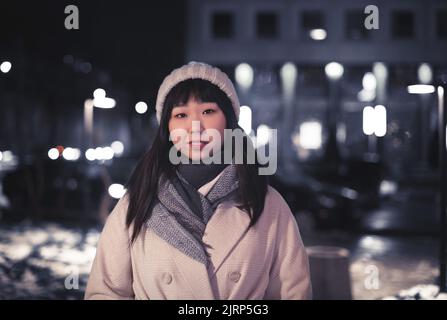 Eine junge Koreanerin in einem warmen Hut auf einer Straße in der Stadt. Ein glückliches sorgloses Mädchen in einem beigen modischen Mantel und an einem Winterabend auf der Straße stehend Stockfoto