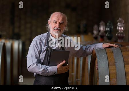 Ein im Kaukasus erfahrener Winzer überprüft eine Weinalterung in Fässern mit einem Touchpad-Tablet. Traditionelles und modernes Technologiekonzept. Stockfoto