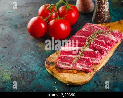 Fleischsteaks auf einem Schneidebrett mit einem Zweig Rosmarin zum Kochen bereit, reife Tomaten auf dunklem Hintergrund. Köstliche hausgemachte Fleischrezepte. Stockfoto