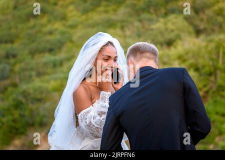 Eine schwarze Braut nimmt Glückwünsche am Telefon entgegen und schaut glücklich auf ihren weißen Ehemann Stockfoto