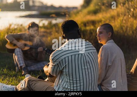 Eine multikulturelle Gruppe von Freunden, die in der Natur am Fluss einen angenehmen Sonnenuntergang genießen, während sie auf dem Gras sitzen und miteinander reden. Ein junger bärtiger Mann, der Gitarre spielt. Hochwertige Fotos Stockfoto
