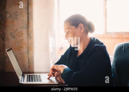 Ernsthafte reife Frau mit Laptop-Computer, während sie zu Hause arbeiten Stockfoto
