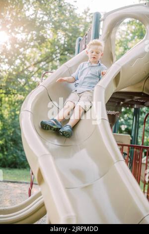 Der blonde 5-jährige Junge rutscht bei Sonnenuntergang eine Rutsche auf dem Spielplatz hinunter. Stockfoto