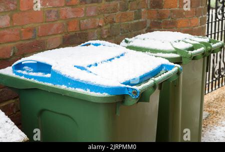 Grüne und blaue Mülltonnen in einem Garten im Winter, Großbritannien. Mit Schnee bedeckte Mülldeckel. Stockfoto