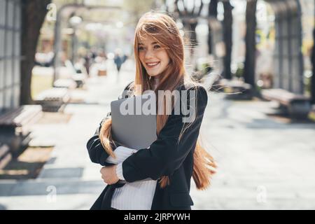 Lächelnde Geschäftsfrau, die die Kamera anschaut, während sie mit einem Laptop im Park spazieren geht. Unternehmenstechnologie. Unternehmensporträt. Stockfoto