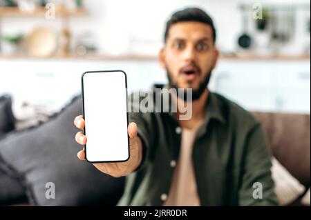 Defokussierter schockierter indischer oder arabischer Mann in Freizeitkleidung, sitzt auf dem Sofa im Wohnzimmer, zeigt Smartphone mit leerem weißen Mock-up-Bildschirm, Platz für Werbung oder Präsentation, schaut erstaunt auf die Kamera Stockfoto