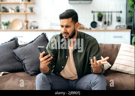 Enttäuschung verwirrt gemischter junger Mann in Freizeitkleidung, sitzt auf dem Sofa im Wohnzimmer, nutzt Mobiltelefon, schreibt online, stöbert in sozialen Netzwerken, liest Nachrichten, Betrachtet den Bildschirm verwirrt, fassungslos Stockfoto