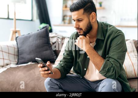 Gadgets-Technologie. Verwirrten indischen oder arabischen Kerl in Freizeitkleidung, sitzt auf dem Sofa in einem Wohnzimmer, mit Smartphone, SMS online, in sozialen Netzwerken surfen, Nachrichten lesen, Sieht den Bildschirm verwirrt an Stockfoto