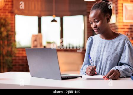 Frau, die mit einem Laptop studiert und sich Notizen im Notebook gemacht hat. afroamerikanische Studentin, die an einer abgelegenen Universität studiert, sieht sich Lerntutorial an und lernt mit Online-Kursen im Internet Stockfoto