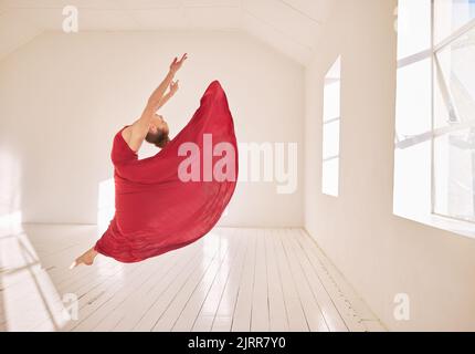 Frau, Ballett und Tanz einer Studentin, die in einem roten Kleid in einem Studiomockup mit weißen Wänden und Sonnenlicht tanzt. Junge professionelle weibliche Ballerina Stockfoto