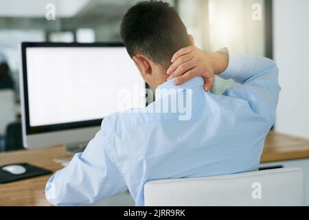 Das Ergebnis einer schlechten Körperhaltung. Rückansicht eines jungen Geschäftsmanns, der seinen Hals in der Hand hielt, während er an seinem Schreibtisch saß. Stockfoto