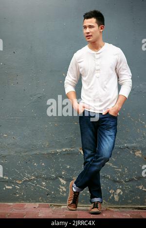 Ein hübscher junger Mann, der mit den Händen in den Taschen an einer grauen Wand steht. Stockfoto