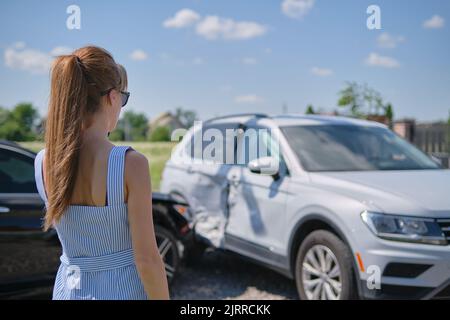 Traurige junge Fahrerin, die in der Nähe ihres zertrümmerten Autos steht und schockiert auf abgestürzte Fahrzeuge bei einem Verkehrsunfall aussieht Stockfoto