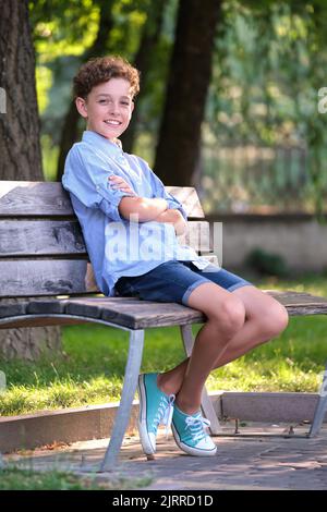 Junge glücklich Kind Junge entspannen auf der Bank im Sommer Park sitzen. Positives Kind, das die Sommerzeit im Freien genießt. Konzept für das Wohlbefinden von Kindern Stockfoto