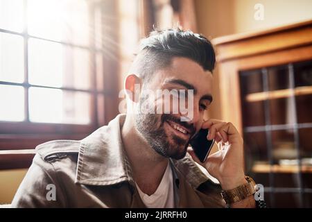 Es ist toll, Ihre Stimme wieder zu hören. Ein hübscher junger Mann lächelt und spricht zu Hause auf einem Handy. Stockfoto