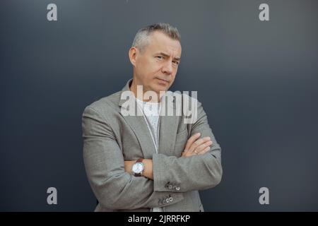 Porträt eines erfolgreichen gealterten grauhaarigen Geschäftsmannes, der mit gekreuzten Armen in stilvollem Anzug und Luxusuhr auf grauem Hintergrund steht. Urbane Lebensart Stockfoto