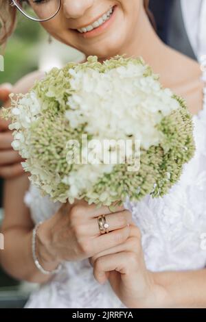 Nahaufnahme vertikaler Hochzeitsstrauß aus weißen und grünen Feldblumen in zugeschnittenen, nicht erkennbaren lächelnden weiblichen Brauthänden Stockfoto