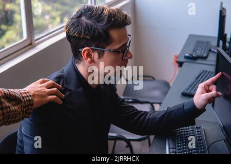 Im Büro arbeitet ein Mann an seinem Computer und sein Chef greift ihn an der Schulter. Kaukasischer blonder Mann am Computer, der auf den Bildschirm zeigt, damit sein Chef etwas sieht. Büromann. Hochwertige Fotos Stockfoto
