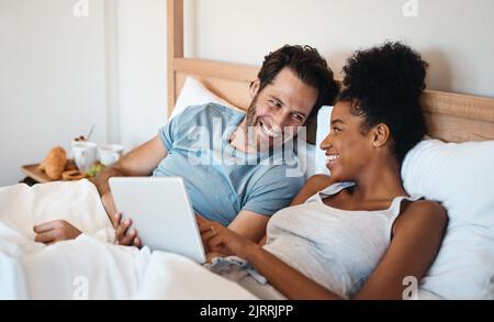 Lachend verheiratetes Paar mit einem Tablet im Bett, das sich am Wochenende zuhause einen lustigen Comedy-Film oder Film ansah. Liebevoll, lächelnd und romantisch Stockfoto