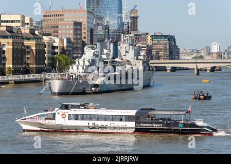 Uber Boat by Thames Clippers River Bus nach Verlassen des Tower Pier in der Nähe der Tower Bridge dreht sich die HMS Belfast im Pool von London auf der Themse Stockfoto