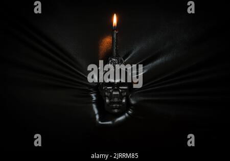 Schwarze Kerze mit Flamme auf Schädel, die von Hand durch schwarzen Stoffhintergrund gedrückt wird. Halloween-Festival-Konzept. Stockfoto