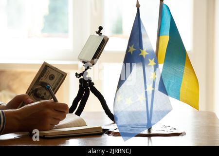 Ein Mädchen mit Dollars in den Händen spricht am Tisch in einem Video-Chat am Telefon, Finanzen und Erfolg, die Flagge der Ukraine und der Europäischen Union Stockfoto