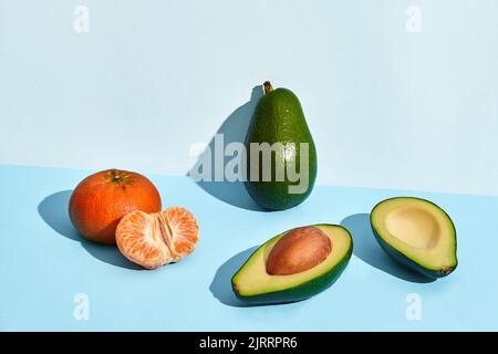 Zusammensetzung aus frischen Früchten, Mandarine, ganzer Avocado und zwei Hälften ausgeschnittenem Avocado auf zweifarbigem Hintergrund Stockfoto