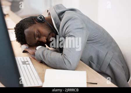 Müde CRM-Callcenter-Mitarbeiter schlafen im Büro auf dem Computertisch mit eingeschaltetes Headset. Erschöpfter junger Geschäftsmann, der ein Nickerchen auf dem Tisch macht. Afrikanisch Stockfoto