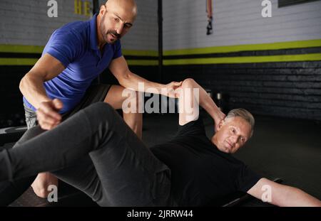 Männlicher Personal Trainer, der einem älteren älteren Mann dabei hilft, Crossover-Bauchmusgeln zu machen. Arbeiten an Kernkraft und Gleichgewicht. Trainieren Sie im Fitnessstudio zu Hause. S Stockfoto