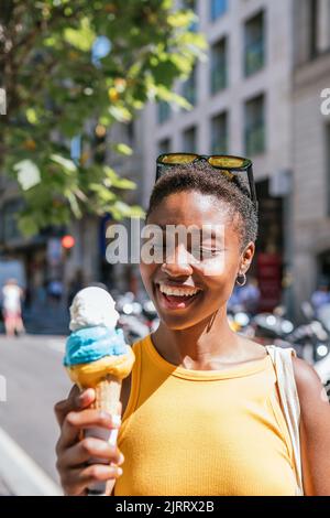 Fröhliche afroamerikanische Frau lächelt, während sie ihr Eis anschaut. Sie ist an einem Sommertag auf der Straße Stockfoto