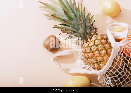 Verschiedene tropische Früchte im Netzbeutel auf beigem Hintergrund. Ananas, Kiwi, Zitronen und Orangen. Draufsicht, flach liegend, Kopierbereich. Stockfoto