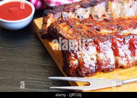 Gegrillte Schweineribs auf einem Holzschneidebrett mit Küchengabel für Fleisch- und Tomatenketchup Stockfoto