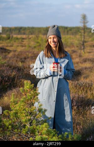 Eine junge Frau mit Hipsterhut und Denim-Mantel geht an Herbstbäumen vorbei und schaut auf den Sonnenuntergang Stockfoto