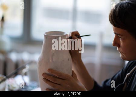 Fokussierte Brünette Frau malt Text mit Pinsel auf weiße Keramikvase am Tisch auf verschwommenem Hintergrund Stockfoto