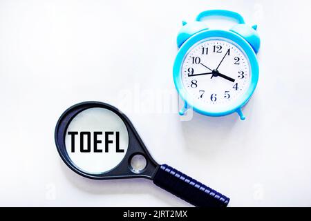 TOEFL ist ein Test von Englisch als Fremdsprache. Text auf einer Lupe auf hellweißem Hintergrund. Im Hintergrund ist eine Uhr. Stockfoto