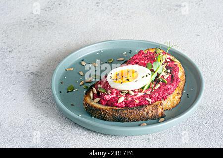 Vollkorn-Toast mit Rübenhummus, Samen, Ei und Mikrogemüse (gekeimt Erbsensprossen) auf hellgrauem Hintergrund. Das Konzept der gesunden Ernährung.. Stockfoto