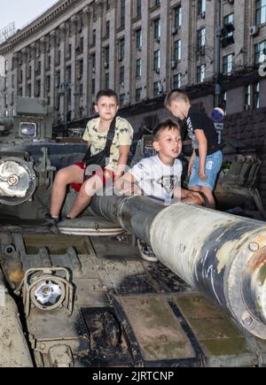 KIEW, UKRAINE - 24. August 2022: Die Jungen spielen auf einem ausgebrannten russischen Panzer. Der Unabhängigkeitstag der Ukraine. Auf Chreschtschatyk in Kiew wird eine Ausstellung zerstörter russischer Waffen organisiert Stockfoto