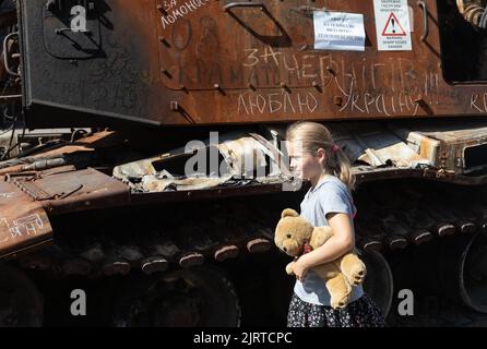 KIEW, UKRAINE - 24. August 2022: Ein Mädchen mit einem Spielzeugbären kommt an einem ausgebrannten russischen Panzer vorbei, der mit der Aufschrift für Tscherniw - I love Ukraine versehen ist. Der Unabhängigkeitstag der Ukraine Stockfoto