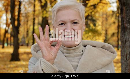 Glückliche ältere Frau freudig lächelnd stehend im Herbstpark reife Großmutter Blick auf die Kamera zeigt Geste okay Symbol der Zufriedenheit Stockfoto