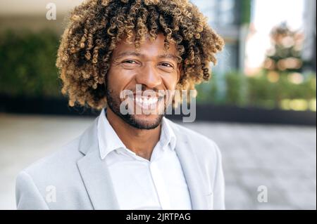 Nahaufnahme eines stolzen, charismatischen, selbstbewussten, lockigen brasilianers oder hispanic-Mannes, erfolgreicher Manager, der im Freien steht, die Kamera anschaut und freundlich lächelt Stockfoto