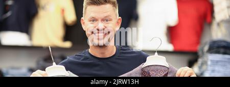 Lächelnder Mann hält zwei Anzüge an Kleiderbügeln im Laden Stockfoto