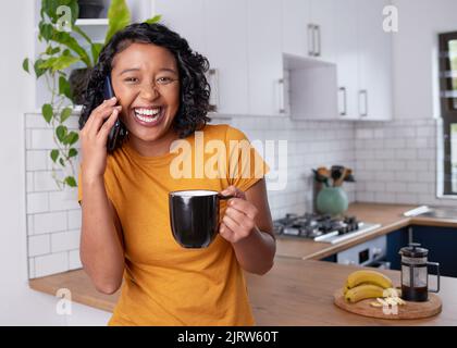 Eine junge, multiethnische Frau lacht, während sie in der Küche telefoniert Stockfoto