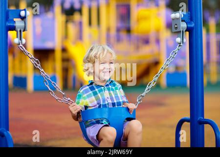 Kinder spielen auf dem Spielplatz im Freien bei Regen. Kinder spielen auf der Schule oder im Kindergarten. Aktives Kind auf farbenfroher Schaukel. Stockfoto