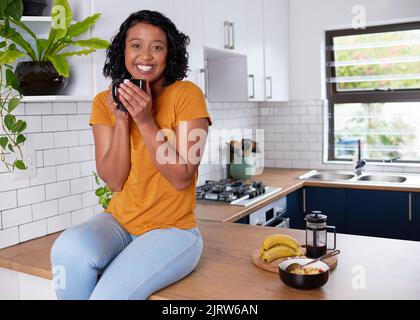 Eine junge, multiethnische Frau sitzt auf ihrer Küchenarbeitsfläche und genießt das Frühstück Stockfoto