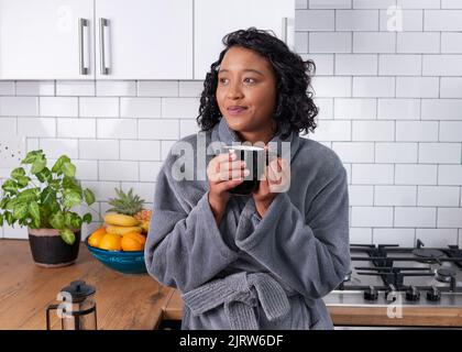 Eine junge Frau genießt es, einen Moment aufzuwachen, während sie in der Küche Kaffee trinkt Stockfoto