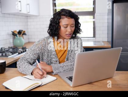 Eine junge, multiethnische Frau arbeitet von zu Hause aus in der Küche und notiert sich Stockfoto